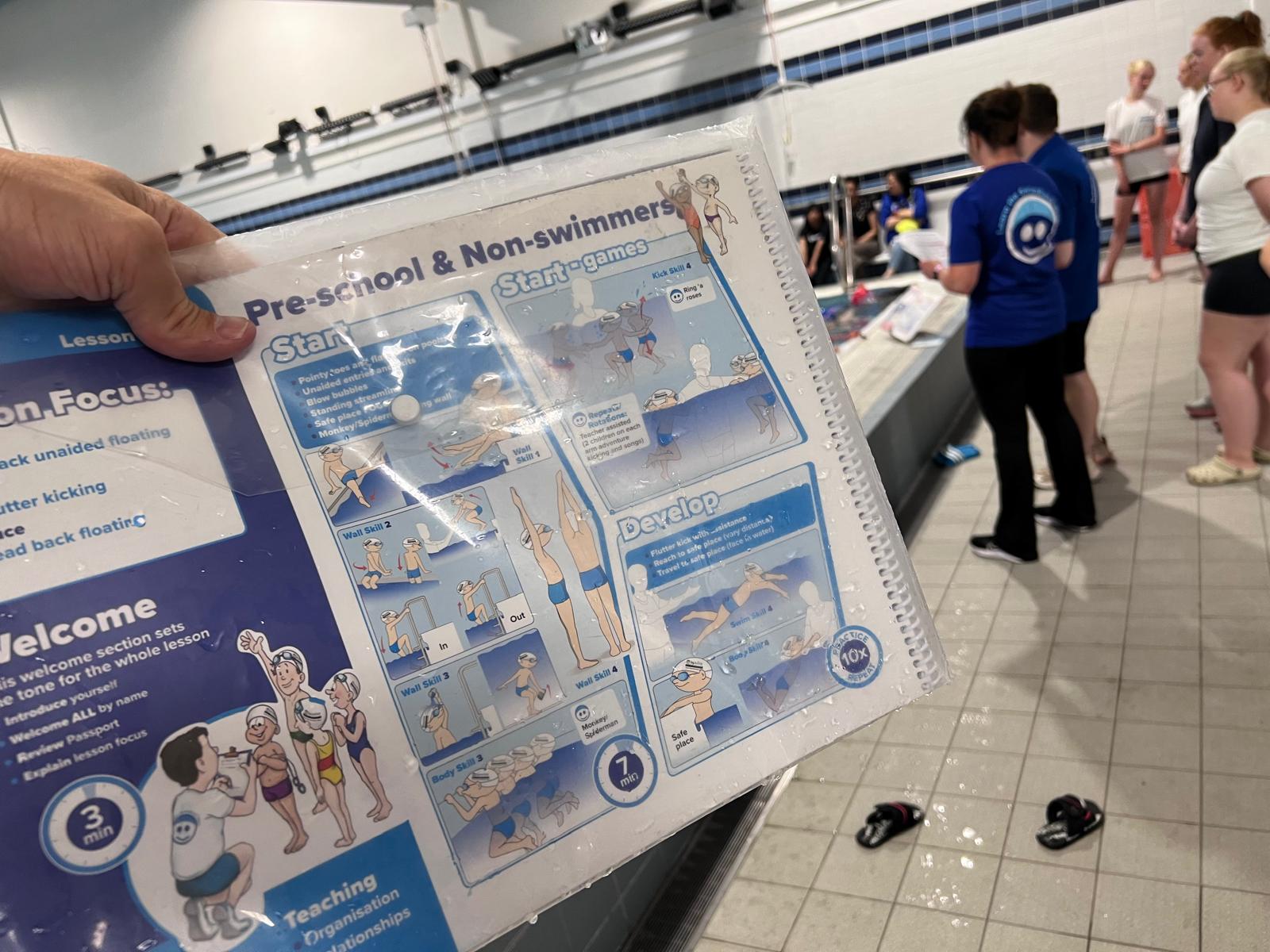 Close-up of a waterproof SwimEasy teacher curriculum passport guide poolside, detailing structured lessons for pre-school and non-swimmers